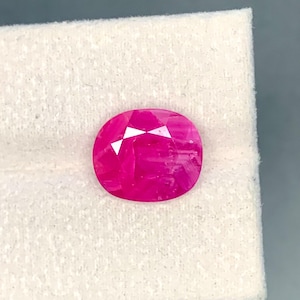 May include: A faceted, oval-shaped ruby gemstone. The gem is a vibrant, deep pink color with a polished surface. The facets reflect light, creating a sparkling effect. The ruby is set against a white background.