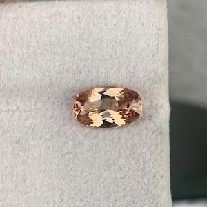 May include: A faceted, oval-shaped gemstone with a peach-colored hue. The gem has a polished surface with multiple facets that reflect light, creating a sparkling effect. The gemstone is set against a white background.