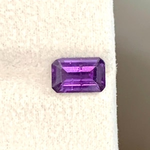 Pode incluir: Uma pedra preciosa de ametista retangular e facetada, com um tom roxo profundo. A gema é cortada com um design de corte em degraus, mostrando sua estrutura interna e brilho. A pedra está sobre um fundo branco.