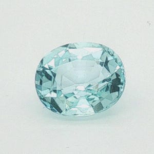 Può includere: Una pietra preziosa a taglio ovale con un colore blu-verde chiaro e una superficie sfaccettata. La pietra presenta una disposizione simmetrica di sfaccettature triangolari e trapezoidali che riflettono la luce, creando un aspetto trasparente.
