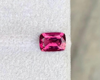 May include: A cushion-cut, vibrant pink gemstone. The gem has a rectangular shape with rounded corners and facets that reflect light. The stone is set against a white background, highlighting its color and clarity. This gemstone is suitable for jewelry.