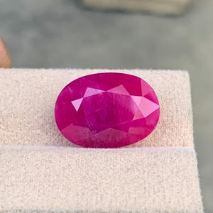 Puede incluir: Una gema de corte ovalado vibrante con un tono rosa magenta intenso y una superficie facetada. La piedra muestra un acabado claro y pulido con múltiples facetas geométricas que reflejan la luz, destacando su color saturado y transparencia.