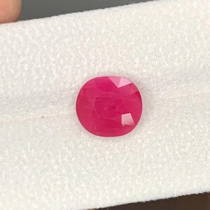 May include: A faceted, oval-shaped ruby gemstone. The gem is a vibrant, deep red color and appears to be set against a white, textured background. The cut of the stone creates multiple reflective surfaces.