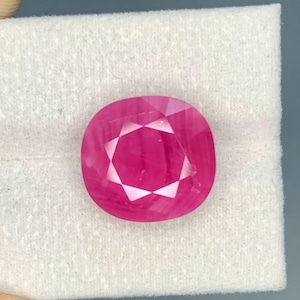 May include: A faceted, cushion-cut ruby gemstone. The gem is a vibrant pinkish-red colour with a starburst pattern in the centre. It is set against a white background.