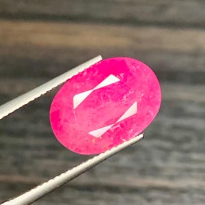 Op de afbeelding: Een levendige, ovaal geslepen, felroze edelsteen wordt vastgehouden door een zilveren pincet. De edelsteen heeft twee witte, geometrische facetten. De achtergrond is een vage, donkere houtnerf.