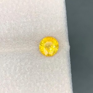 Puede incluir: Una gema amarilla redonda y facetada, posiblemente un zafiro o citrino, sobre un fondo de tela blanca. La gema es de un color amarillo vibrante y soleado, con un corte brillante que captura la luz.