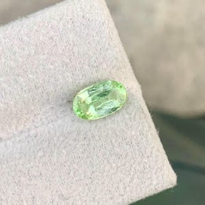 Puede incluir: Una gema ovalada de color verde claro con una superficie pulida. La gema está colocada sobre un fondo blanco texturizado. El color de la piedra es un verde translúcido claro, con reflejos internos.