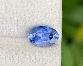 Puede incluir: Gema de zafiro ovalada y facetada. El zafiro es de un color azul profundo y vibrante con inclusiones internas. La gema está sobre un fondo blanco, con un fondo verde borroso.