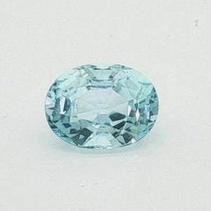 May include: A loose oval-cut gemstone with a light blue colour and a faceted surface. The stone features a symmetrical arrangement of triangular and trapezoidal facets that reflect light, creating a clear, transparent appearance with internal depth.