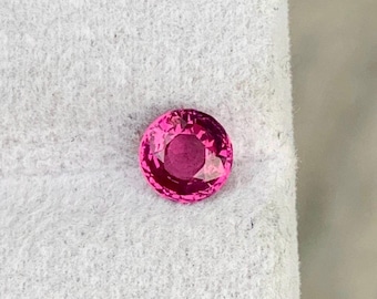 Puede incluir: Una gema redonda y facetada en un tono rosa vibrante. La gema se encuentra sobre un fondo blanco texturizado. El corte de la gema permite que la luz se refleje, creando un efecto brillante. Adecuado para la fabricación de joyas.
