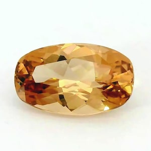 Könnte beinhalten: Ein oval geschliffener Edelstein mit einem warmen, gold-orangenen Farbton. Der Edelstein hat mehrere Facetten, die das Licht reflektieren und einen funkelnden Effekt erzeugen. Der Stein ist vor einem schlichten weißen Hintergrund platziert.