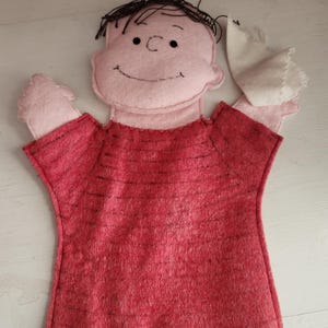May include: A felt hand puppet featuring a light pink face with black embroidered facial features and dark brown yarn hair. The puppet wears a red shirt with horizontal black stripes and has a small white fabric piece attached to one hand.