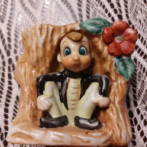 May include: A ceramic figurine of a small elf sitting in a tree stump with a red flower. The elf is wearing a black jacket, white pants, and black shoes. The tree stump is brown and has a white base.