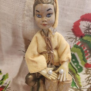 May include: A vintage figurine of a person playing drums. The figure has a yellow outfit, a gold head covering, and blue eyes. The hands hold two white pearls. The drums are beige and the figure is seated.