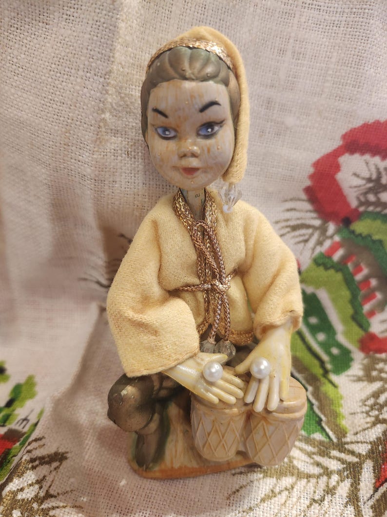 Op de afbeelding: Een vintage beeldje van een persoon die drums speelt. De figuur heeft een gele outfit, een gouden hoofdbedekking en blauwe ogen. De handen houden twee witte parels vast. De drums zijn beige en de figuur zit.