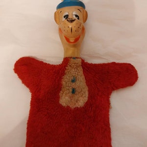 May include: A red fuzzy hand puppet with a brown and blue face, wearing a blue and white hat. The puppet has a wide smile and large eyes.