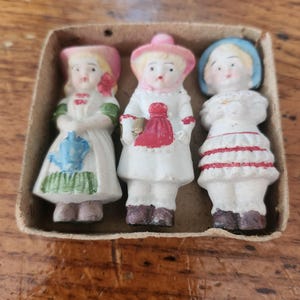 May include: Three antique porcelain figurines of children in a cardboard box. The figurines are dressed in white dresses with colorful accents. The first doll has a pink hat and a blue and green design. The second doll has a pink hat and a red bag. The third doll has a blue hat and red stripes.
