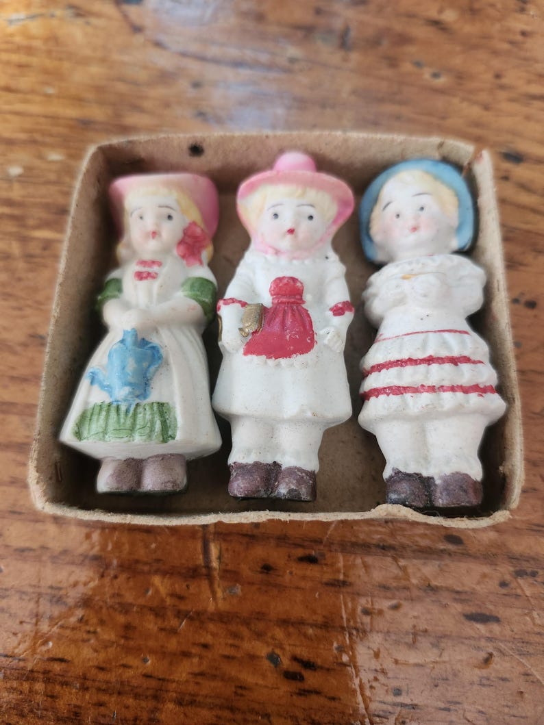May include: Three antique porcelain figurines of children in a cardboard box. The figurines are dressed in white dresses with colorful accents. The first doll has a pink hat and a blue and green design. The second doll has a pink hat and a red bag. The third doll has a blue hat and red stripes.