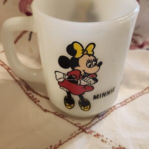 Puede incluir: Taza de cer&aacute;mica blanca con una imagen de dibujos animados de Minnie Mouse con un vestido rojo y un lazo amarillo. La taza tiene la palabra "MINNIE" impresa en la parte inferior.