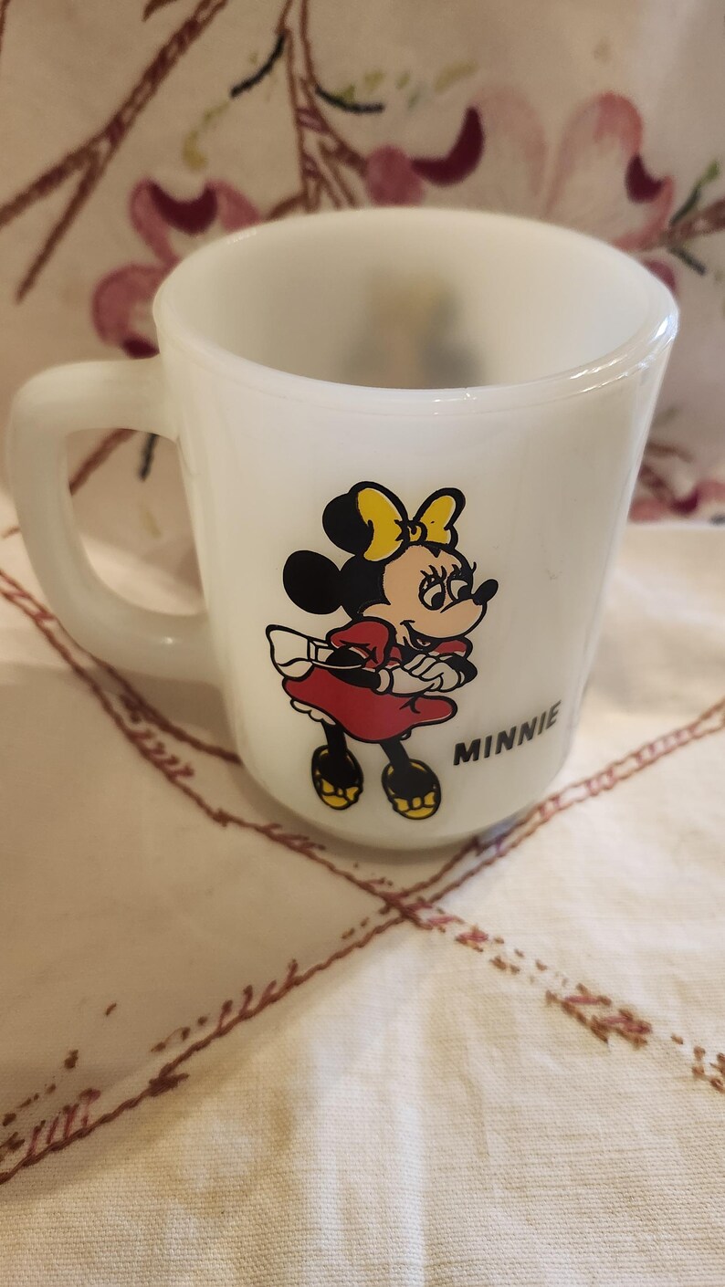 Puede incluir: Taza de cer&aacute;mica blanca con una imagen de dibujos animados de Minnie Mouse con un vestido rojo y un lazo amarillo. La taza tiene la palabra "MINNIE" impresa en la parte inferior.