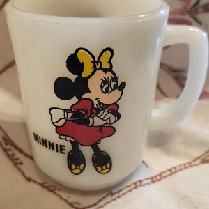 Puede incluir: Taza de cer&aacute;mica blanca con una ilustraci&oacute;n de Minnie Mouse. Minnie lleva un vestido rojo, zapatos amarillos y un lazo amarillo. La taza tiene la palabra "MINNIE" impresa en la parte inferior.