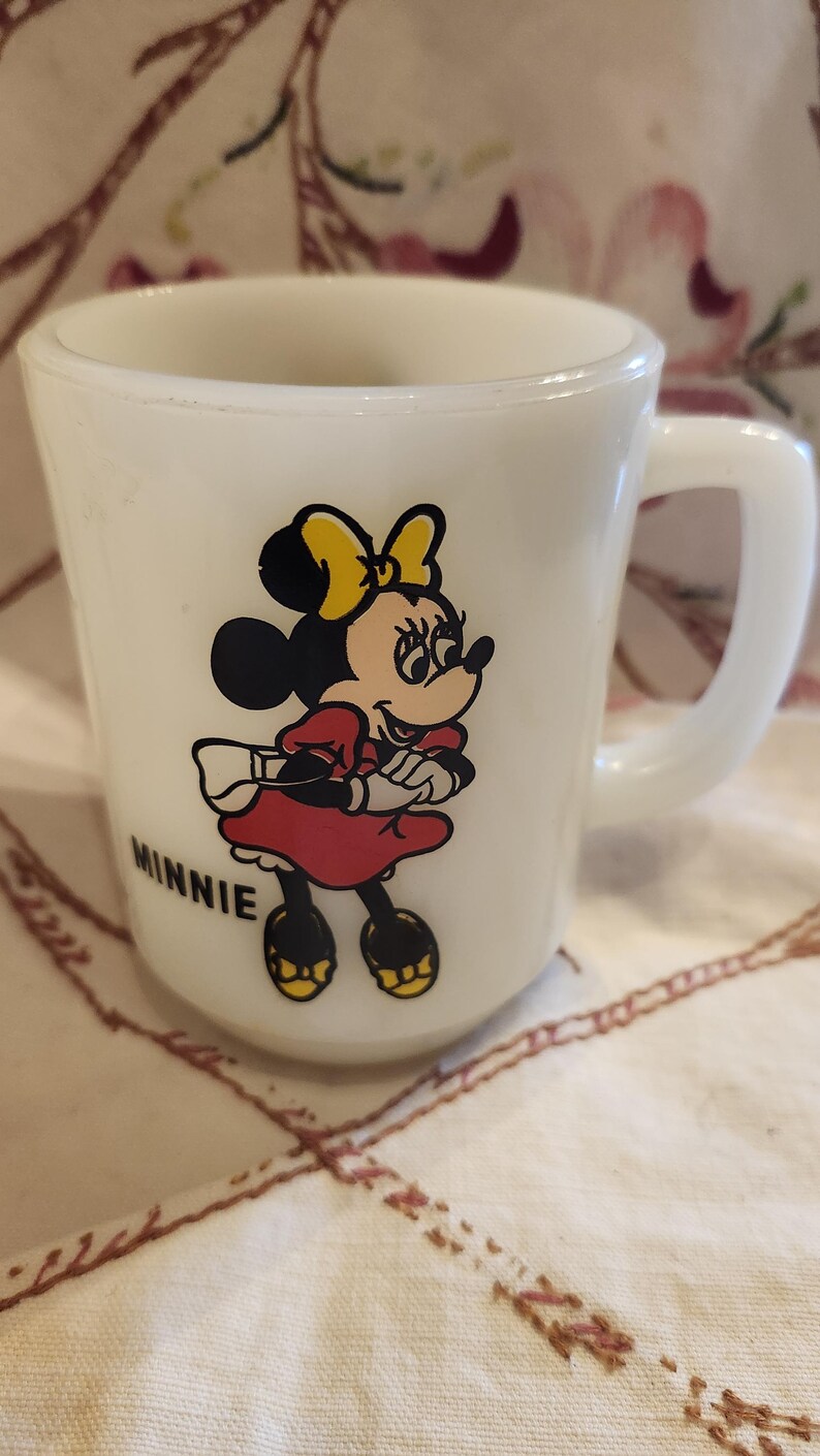 Puede incluir: Taza de cer&aacute;mica blanca con una ilustraci&oacute;n de Minnie Mouse. Minnie lleva un vestido rojo, zapatos amarillos y un lazo amarillo. La taza tiene la palabra "MINNIE" impresa en la parte inferior.