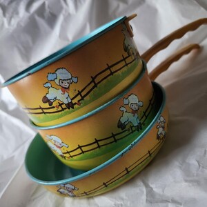 May include: A stack of three round metal pans with yellow and orange gradient exteriors featuring a repeating illustration of white lambs jumping over a black fence. The pans have light blue interior rims and long, curved yellow handles.