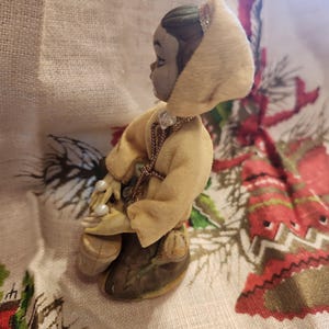 Op de afbeelding: Een klein beeldje van een persoon met een gele jas met capuchon en parels. Het beeldje heeft een grijs gezicht en donker haar. De jas is afgezet met een gouden ketting. Het beeldje knielt op een oppervlak met patronen.