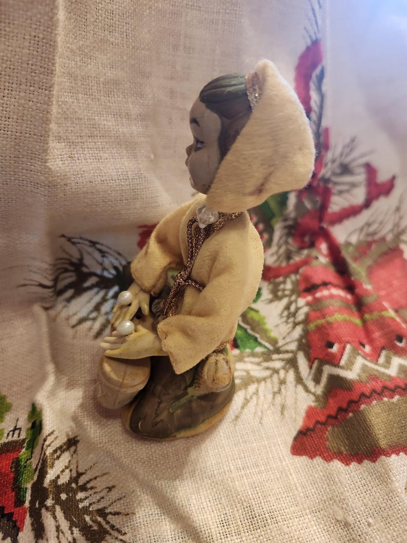 Op de afbeelding: Een klein beeldje van een persoon met een gele jas met capuchon en parels. Het beeldje heeft een grijs gezicht en donker haar. De jas is afgezet met een gouden ketting. Het beeldje knielt op een oppervlak met patronen.