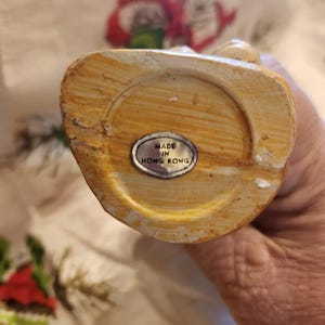 Op de afbeelding: Een beige beeldje met een textuur. De onderkant van het beeldje heeft een cirkelvormige inkeping en een kleine, ovale sticker met de tekst "MADE IN HONG KONG".