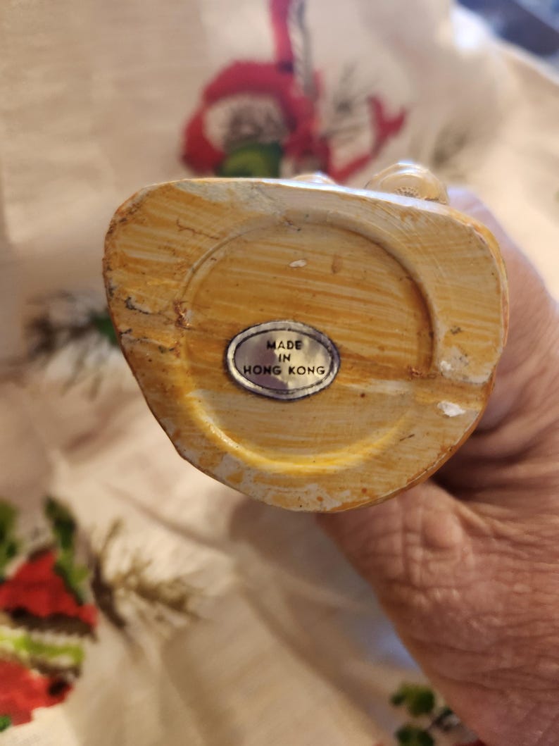 Op de afbeelding: Een beige beeldje met een textuur. De onderkant van het beeldje heeft een cirkelvormige inkeping en een kleine, ovale sticker met de tekst "MADE IN HONG KONG".