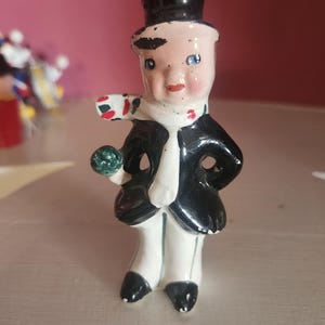 May include: A ceramic figurine of a person wearing a black top hat, a black coat, and white trousers. The figure has a white scarf with red and green accents, blue eyes, and holds a small green spherical object in one hand. The paint shows wear.