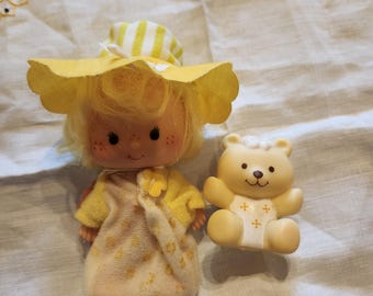 May include: A vintage doll with blonde hair, freckles, and a yellow hat and dress. A small, tan teddy bear with cross details is next to the doll. The doll's dress has a floral pattern, and a green bow is attached.