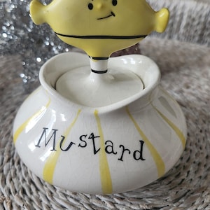 May include: A white ceramic mustard jar with yellow stripes and the word "Mustard" written in black. The lid is a yellow and orange cartoon character with a grumpy expression.