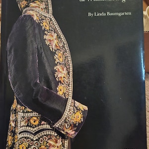 May include: A hardcover book titled Eighteenth-Century Clothing at Williamsburg by Linda Baumgarten. The black cover features a detailed illustration of a dark coat with intricate yellow, pink, and green floral embroidery and trim.