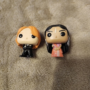 May include: Two Funko Pop vinyl figures. One figure has orange hair and wears a black outfit. The other figure has black hair and wears a pink and white outfit. Both figures have large black eyes and are standing on a textured surface.