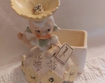 May include: A porcelain figurine of a girl wearing a yellow flower hat and a white dress with yellow flowers. She is holding a tag that says "August".