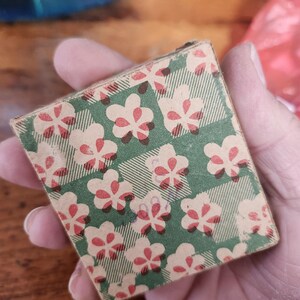 May include: A small, rectangular box with a green and white plaid pattern, overlaid with a repeating floral design in cream and red. The box is held in a hand, with the focus on the patterned surface.