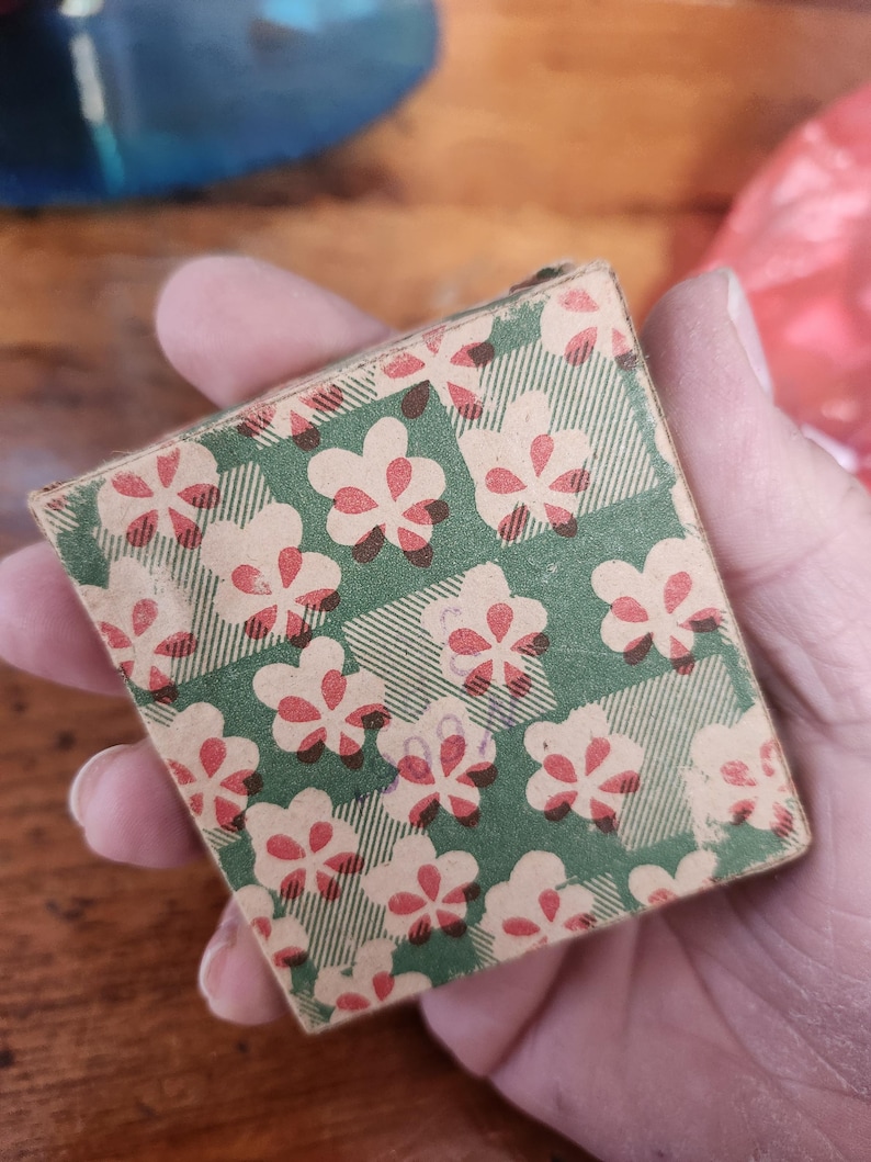 May include: A small, rectangular box with a green and white plaid pattern, overlaid with a repeating floral design in cream and red. The box is held in a hand, with the focus on the patterned surface.
