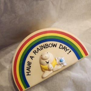 May include: A cream-colored, rainbow-shaped decorative item with the text "Have a Rainbow Day!". It features a cartoon bear wearing a yellow shirt and a small blue bear. The rainbow has red, orange, yellow, green, and blue stripes.