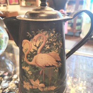 May include: A vintage black metal teapot with a lid and handle. The teapot features a painted design of two pink flamingos amidst green foliage and flowers. The teapot is displayed on a glass surface.
