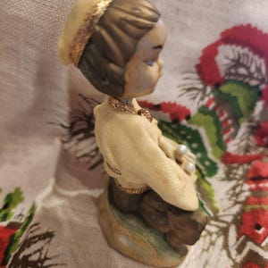Op de afbeelding: Een vintage beeldje van een zittend kind, gekleed in een cr&egrave;mekleurige outfit en een bijpassende hoed met gouden rand. Het beeldje heeft een gouden ketting en houdt parels vast. Het haar van het kind is bruin en het beeldje staat op een voet met groene en bruine tinten.