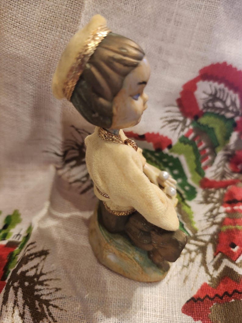 Op de afbeelding: Een vintage beeldje van een zittend kind, gekleed in een cr&egrave;mekleurige outfit en een bijpassende hoed met gouden rand. Het beeldje heeft een gouden ketting en houdt parels vast. Het haar van het kind is bruin en het beeldje staat op een voet met groene en bruine tinten.