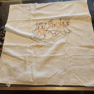 May include: A white square kitchen towel featuring an embroidered design of two kittens sitting on a blue outlined mat. Each kitten wears an orange bow. The text "Friday Clean" is embroidered in orange thread below the kittens on the towel.