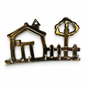 Könnte beinhalten: Ein messingfarbener Wand-Schlüsselhalter aus Metall mit einer ausgeschnittenen Silhouette eines Hauses mit Schornstein, einem Baum und einem Lattenzaun. Die Basis des Designs enthält fünf kleine silberfarbene Metallhaken zum Aufhängen.