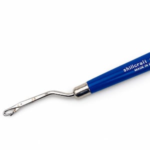 May include: A blue Shillcraft Readicut latch hook tool with a silver metal hook and a smooth handle. The handle features the white printed text "shillcraft readicut" and "MADE IN ENGLAND" on its side, with a metallic ferrule connecting the parts.