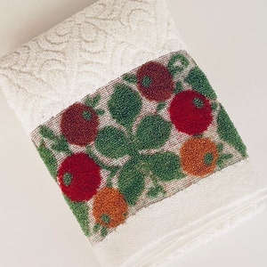May include: Folded white hand towel with a floral design. The towel features a decorative band with red and orange flowers and green leaves. The top portion has a textured pattern.