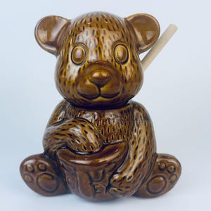 May include: A brown ceramic honey pot shaped like a bear, with a matching honey dipper. The bear is sitting with a small bowl in its paws. The honey pot has a glossy finish and is set against a white background.