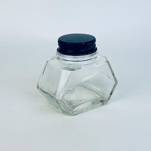 May include: A clear glass inkwell with a faceted design and a black screw-on lid. The inkwell has a geometric shape and is empty. The background is a plain white surface.