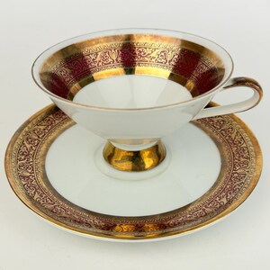 May include: White teacup and saucer set with gold and burgundy accents. The cup and saucer feature ornate patterns and a gold rim. The teacup has a gold pedestal base and handle. A classic tea set.